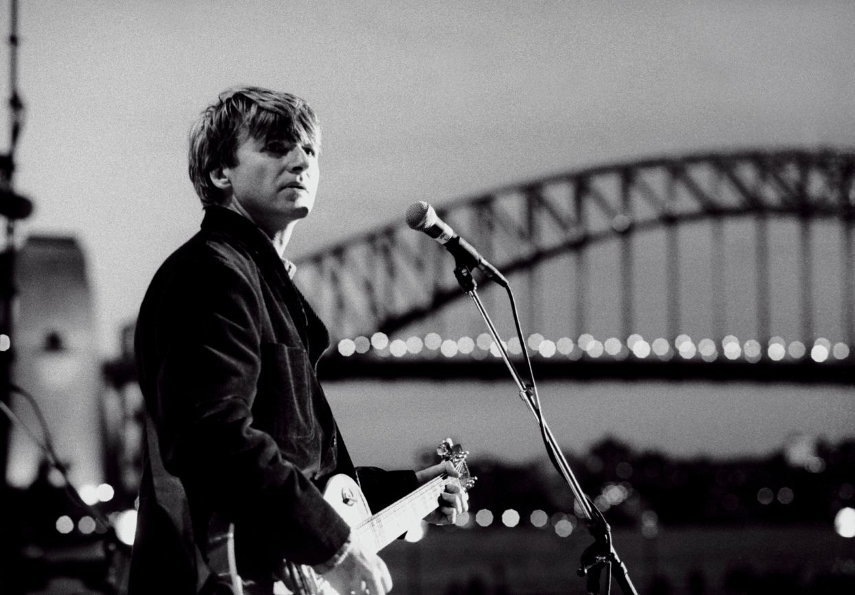 Black and white photo of Neil Finn of Crowded House performing in Sydney with the Harbour Bridge in the background, taken by Tony Mott.