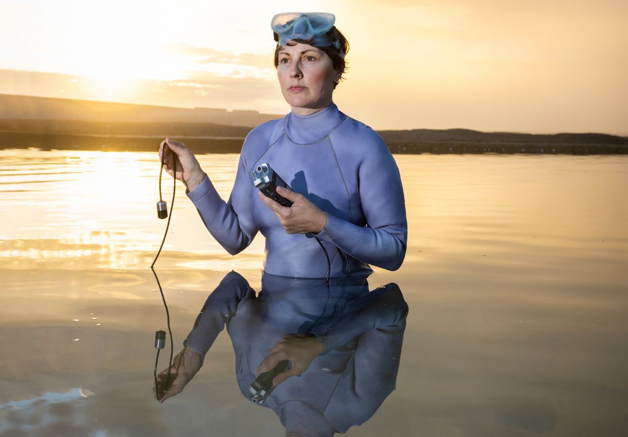 Artistic portrait of a person standing in still water at sunset, wearing a wetsuit and snorkel mask, holding environmental monitoring equipment. 