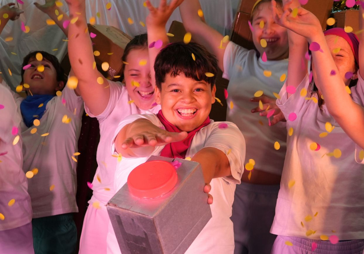 Children in a show throwing colourful confetti while one child presses a large red button on a prop box.