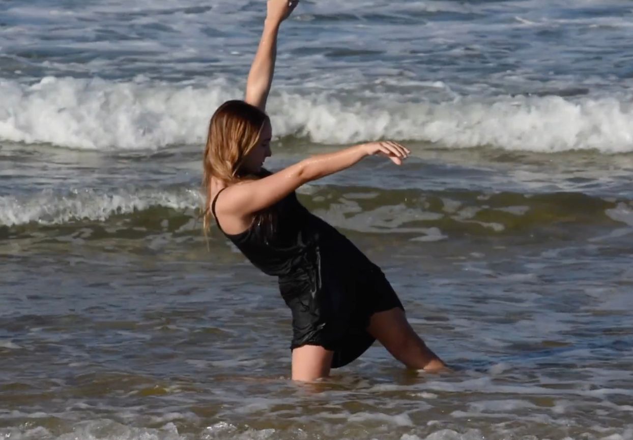 Catapult dancer dressed in black performing in shallow ocean water.