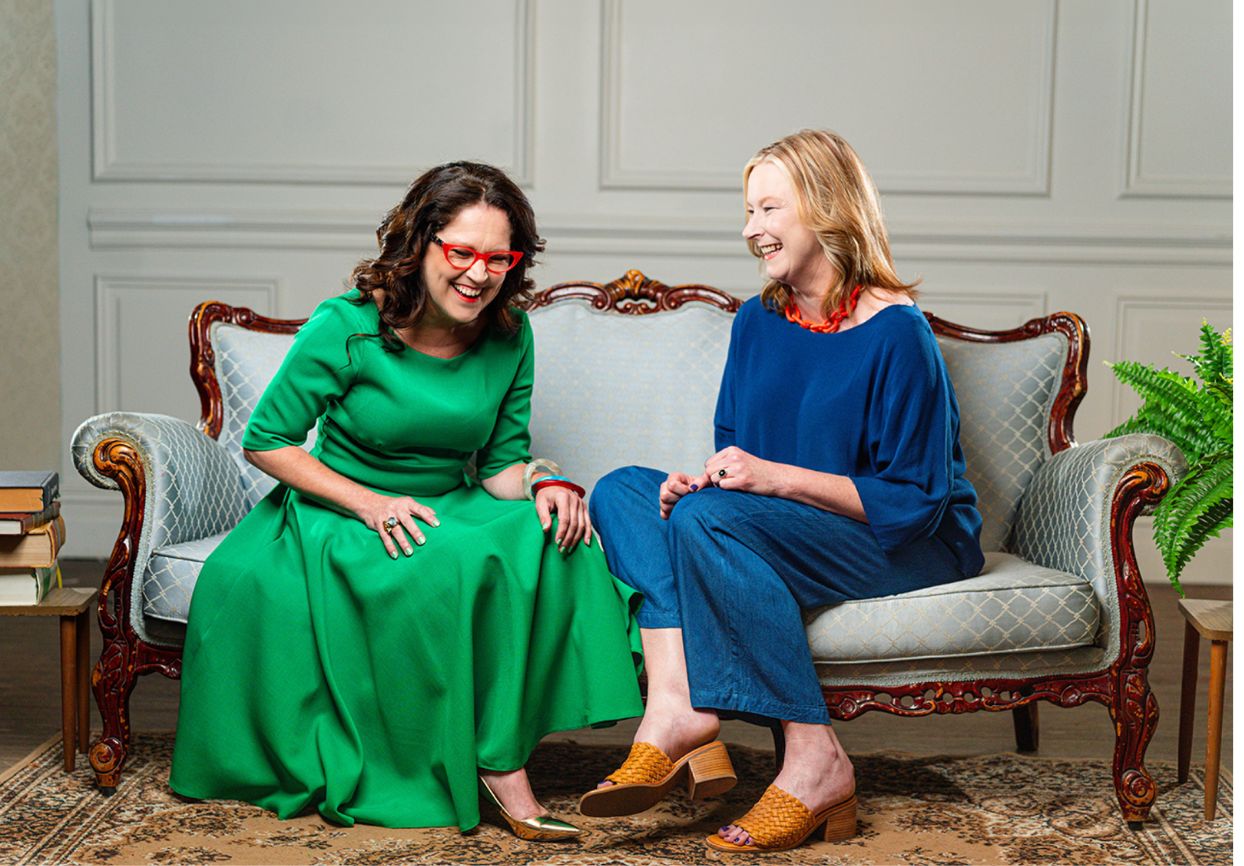 Annabel Crabb in a bright green dress sitting with Leigh Sales in a blue outfit, talking and laughing together on a couch.