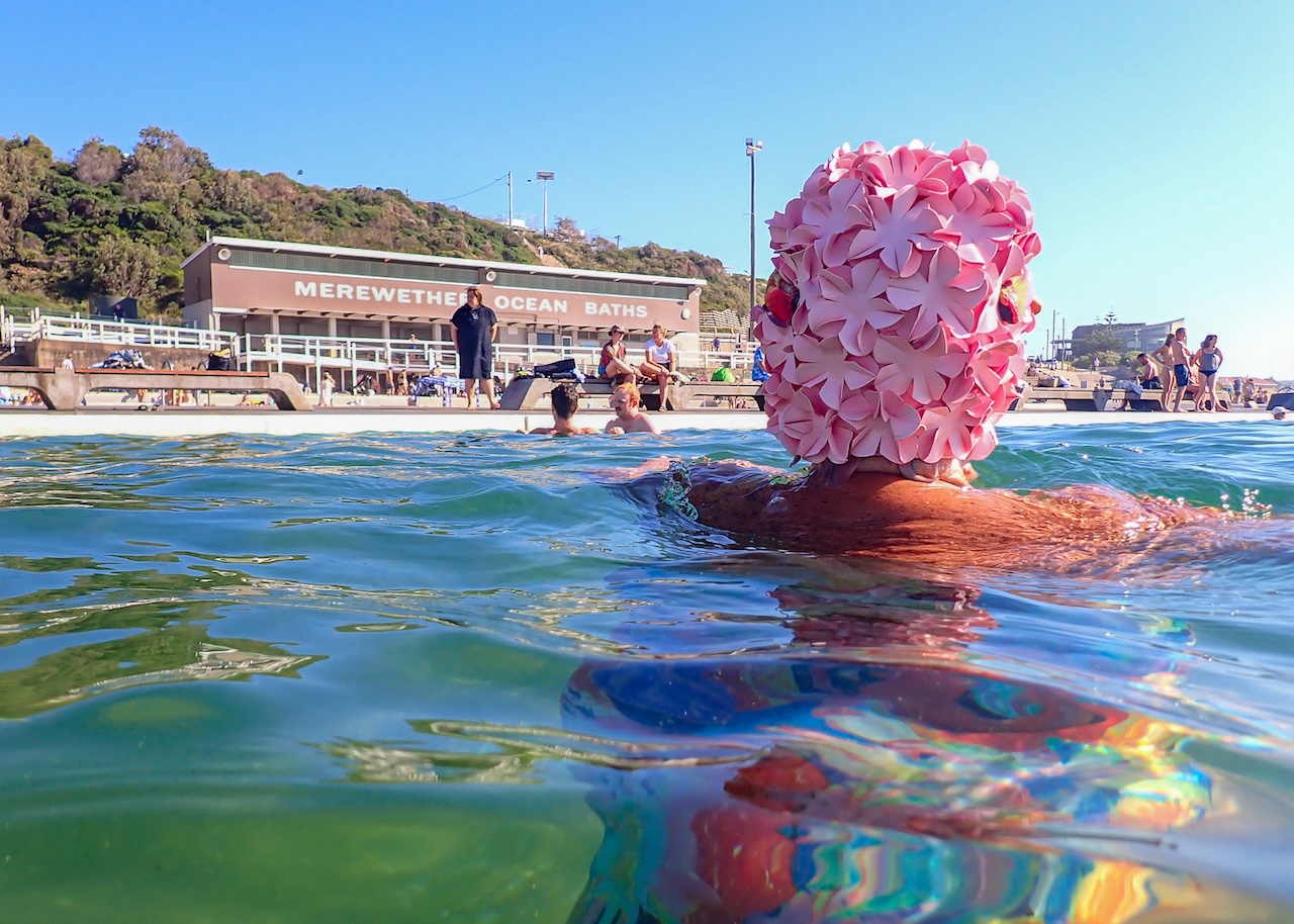 Swimmer wearing a pink flower-covered swim cap in Merewether Ocean Baths, with the pavilion and other swimmers in the background.